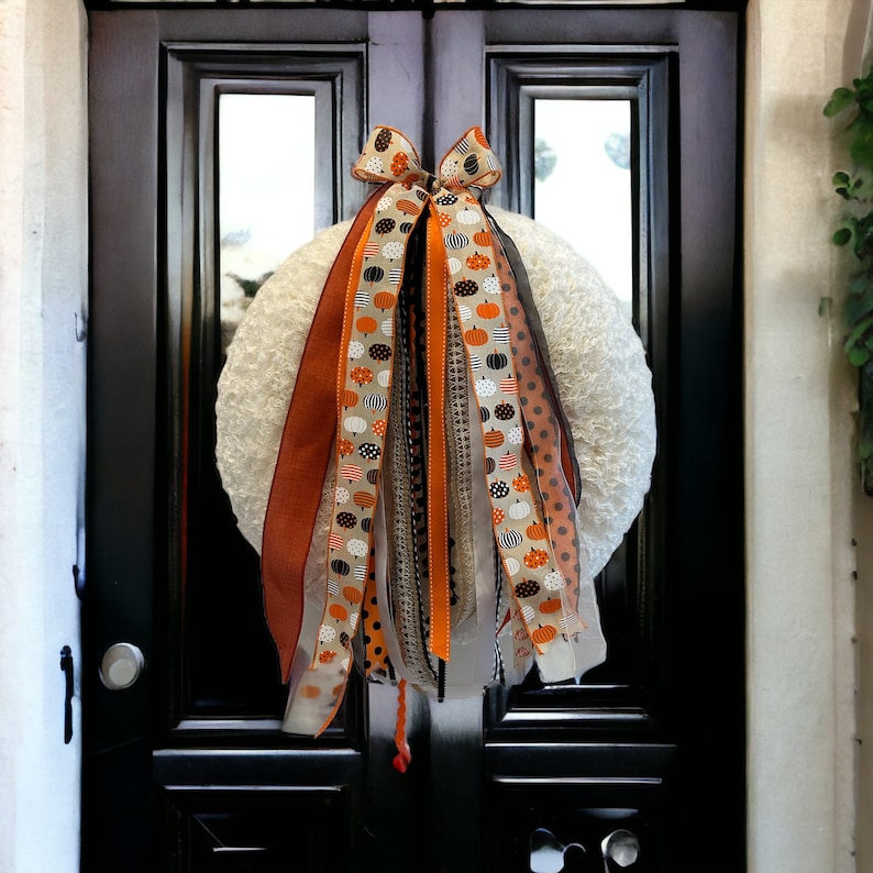 May include: A white wreath with orange, black, and white ribbon streamers with a pumpkin pattern. The wreath is hanging on a black door.