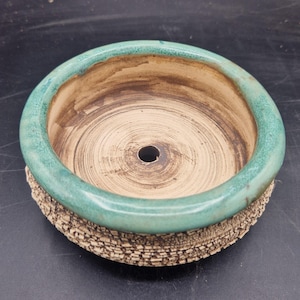 May include: A round ceramic planter with a textured, crackled beige exterior and a smooth, glossy teal green rim. The interior is a light tan colour with visible circular pottery wheel markings and a single drainage hole centred at the base.