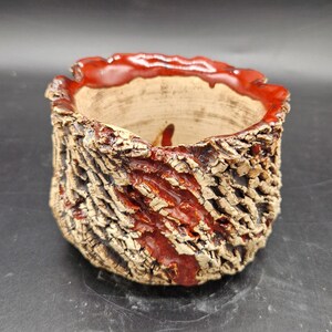 Puede incluir: Una maceta de cerámica hecha a mano con un exterior texturizado similar a la corteza. El borde y una franja vertical están esmaltados en rojo vibrante. El interior es de color beige natural sin esmaltar, creando una estética rústica.