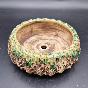 Puede incluir: Una maceta de cerámica hecha a mano con un exterior texturizado en forma de corteza. El borde presenta un esmalte verde y crema, mientras que el interior es marrón natural. La maceta tiene un orificio de drenaje.
