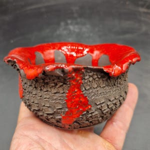 Puede incluir: Un cuenco de cerámica pequeño y redondo con una superficie exterior oscura, rugosa y texturizada. El borde y la parte superior presentan un esmalte rojo brillante que gotea por los lados, contrastando con la textura de arcilla gris oscuro mate y agrietada.