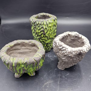 Puede incluir: Tres macetas de cerámica texturizadas en tonos verdes y grises. Las macetas tienen una apariencia rugosa y orgánica, con una maceta alta y cilíndrica y dos macetas más pequeñas en forma de cuenco. Adecuadas para plantas pequeñas o suculentas.