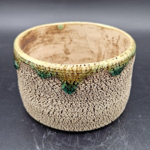 Puede incluir: Un cuenco de cerámica hecho a mano con un exterior beige texturizado. El borde está adornado con un ribete verde y amarillo, y el interior es de color marrón claro. El cuenco tiene forma redonda y es adecuado para diversos usos.