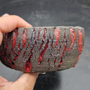 Può includere: Una ciotola in ceramica circolare con un esterno scuro, ruvido e color carbone, caratterizzato da profonde fessure verticali. Uno smalto rosso brillante cola attraverso queste crepe, creando un forte contrasto con la superficie opaca.