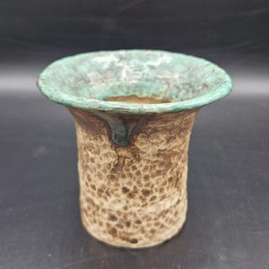 Könnte beinhalten: Eine handgefertigte Keramikvase mit einer strukturierten, gesprenkelten Oberfläche in Braun- und Beigetönen. Der Rand hat eine kontrastierende türkisgrüne Farbe. Die Vase hat eine ausgestellte Öffnung und einen zylindrischen Körper.