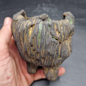 May include: A small, hand-built ceramic pot with a rough, dark charcoal-coloured exterior featuring deep vertical grooves and textured ridges. The crevices reveal hints of bright green and earthy yellow glaze, and the pot stands on three short feet.