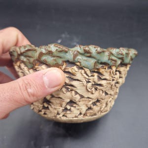Puede incluir: Un pequeño cuenco de cerámica hecho a mano con un exterior texturizado y un borde ondulado. El cuenco presenta una base marrón claro con un patrón detallado y un borde esmaltado turquesa y marrón. El cuenco es sostenido por una mano.