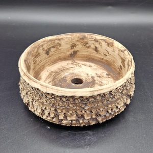 Puede incluir: Una maceta redonda de cerámica hecha a mano con un exterior texturizado similar a la corteza. La maceta es de color marrón claro con un borde y un interior marrón más oscuro. Cuenta con un orificio de drenaje, ideal para plantas pequeñas.