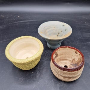 Könnte beinhalten: Drei kleine Keramiktöpfe mit Abflusslöchern. Einer ist gelb texturiert, einer hellblau mit dunklen Sprenkeln und einem ausgestellten Rand, und einer beige mit horizontalen Rillen und einem dunkelrot glasierten Rand.
