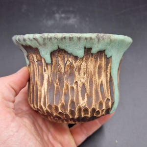 Puede incluir: Una maceta de cerámica hecha a mano con un exterior texturizado y tallado. La maceta tiene una base marrón con un esmalte verde claro que gotea sobre el borde. La maceta se sostiene en una mano, mostrando su tamaño y artesanía. Ideal para plantas pequeñas o como pieza decorativa.
