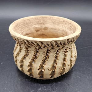 Puede incluir: Un cuenco de cerámica beige con un diseño texturizado y tallado. El cuenco tiene una abertura ancha y una base redondeada. El exterior presenta un patrón repetitivo de líneas y ranuras diagonales, creando una estética rústica.