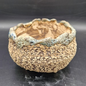 Puede incluir: Una pequeña maceta redonda de cerámica hecha a mano con una superficie texturizada y agrietada. La maceta tiene una base marrón claro con un borde azul-verdoso. El interior es de color marrón oscuro. Esta maceta decorativa es adecuada para plantas pequeñas.