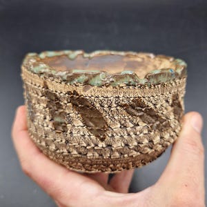 Puede incluir: Un cuenco de cerámica hecho a mano con un patrón texturizado similar al tejido en tonos marrones y beige. El borde presenta un esmalte turquesa, añadiendo un toque de color. El exterior del cuenco tiene una superficie táctil única.