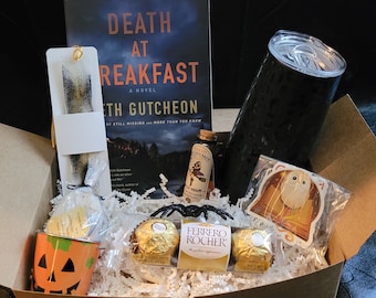 May include: A brown cardboard box filled with a variety of items, including a book titled "Death at Breakfast", a black travel mug, a small bottle of liquid, a package of chocolate candies, a pumpkin-shaped container, a bookmark, and a sticker with an owl design.