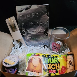 May include: A brown cardboard box filled with a book titled "Tin Foil Butterfly", a glass with a sugar skull design, a bookmark, a small container of coffee, a bag of sour candy, and a small bottle of liquid.