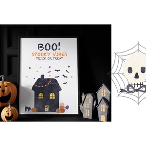 May include: A framed white poster featuring the text BOO! SPOOKY VIBES TRICK OR TREAT, a dark house, bats, and pumpkins. Beside it is a graphic of a white skull centered on a gray spider web with a black bow. Two orange jack-o'-lanterns sit nearby.