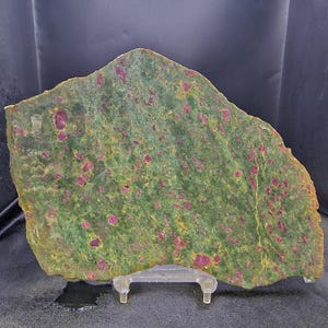 May include: A flat, irregularly shaped slab of zoisite mineral featuring a mottled green and yellow base embedded with numerous small, vibrant magenta-pink ruby crystals throughout the surface, supported by a clear plastic display stand.