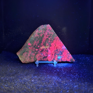 May include: A triangular-shaped rock with a vibrant pink and green pattern, displayed on a clear stand. The rock's surface has a speckled texture, with the pink areas appearing to glow under a blue light.