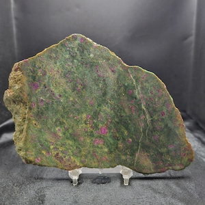 May include: A flat, irregularly shaped slab of zoisite mineral featuring a mottled dark green base color with numerous embedded, vibrant magenta-colored ruby crystals scattered throughout the surface, supported by a clear plastic display stand.