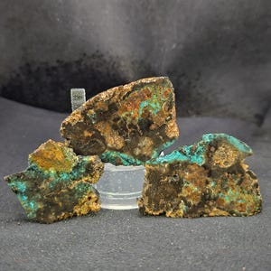 May include: Three rough-cut mineral specimens with a mix of brown, teal, and gold colors. The mineral pieces are displayed on a clear plastic stand against a dark background. The textures are rough and uneven.
