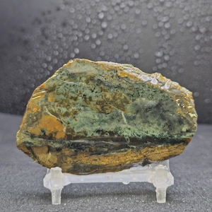 May include: A polished, irregularly shaped stone with a mix of colors. The stone features bands of green, brown, and gold hues. It is displayed on a clear, plastic stand, showcasing its natural textures and patterns.