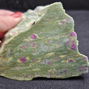 May include: A rough-cut, green stone with multiple, small, bright pink ruby inclusions. The stone has a layered appearance with light yellow and white streaks. The edges are uneven, and the surface is textured. The stone is held by a hand.