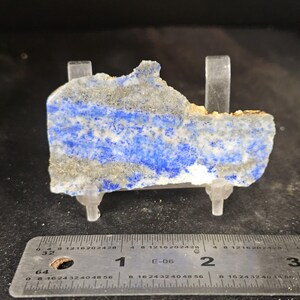 May include: A rectangular slab of lapis lazuli, showcasing a vibrant blue hue with streaks of white and gold. The stone is held by clear plastic supports, with a ruler in inches for scale. The natural stone has a rough, uneven edge.