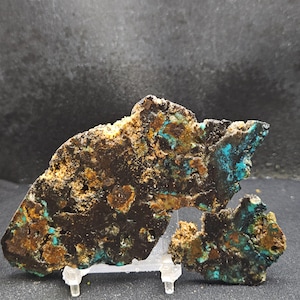 May include: A rough-hewn stone specimen with a dark brown base, accented by patches of turquoise and rust-colored minerals. The stone is displayed on a clear stand, showcasing its natural, irregular shape and texture. The overall appearance suggests a raw, geological sample.