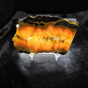 May include: A translucent slab of Bumblebee Jasper mineral specimen featuring vibrant bands of bright orange, yellow, and deep black. The stone is backlit, highlighting its fibrous, radiating crystalline structure and is displayed on a clear stand.