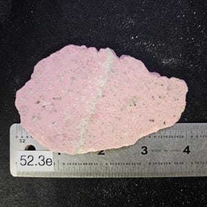 May include: A flat, irregularly shaped slab of pink rhodonite mineral with speckled grey and white inclusions. A thin, light-colored vein runs diagonally across the surface. A metal ruler is positioned underneath, showing a width of 3.5 inches.