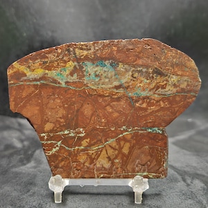 May include: A polished, rectangular stone slab with a rich, reddish-brown base color. The stone features striking veins of turquoise and yellow, creating a natural, abstract pattern. The stone is displayed on a clear, plastic stand.