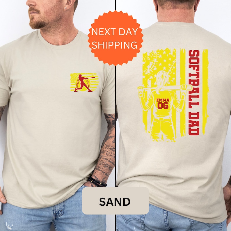 May include: Sand-colored t-shirt with a softball player graphic on the front and a softball player with an American flag design on the back. The back design includes the text "Softball Dad" and "Emma 06".