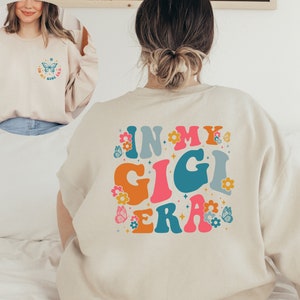 May include: A beige sweatshirt with the text "In My Gigi Era" in colorful retro lettering and a butterfly graphic. The text is surrounded by small flower and butterfly graphics.