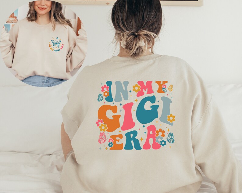 May include: A beige sweatshirt with the text "In My Gigi Era" in colorful retro lettering and a butterfly graphic. The text is surrounded by small flower and butterfly graphics.