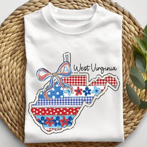 May include: White t-shirt featuring a West Virginia state outline design. The design incorporates a bow and floral elements in red, white, and blue patterns. The words "West Virginia" are written in a script font.