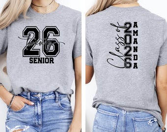 以下が含まれることがあります： 前面に黒で「26 Twenty Senior」と書かれたグレーのTシャツ。Tシャツの背面には、黒で「Class of 2026 Amanda」と書かれています。Tシャツは半袖です。