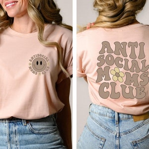 May include: A light pink t-shirt with a brown smiley face and the text "Antisocial Moms Club" on the front. The back of the shirt has the text "Antisocial Moms Club" in brown with a yellow flower.