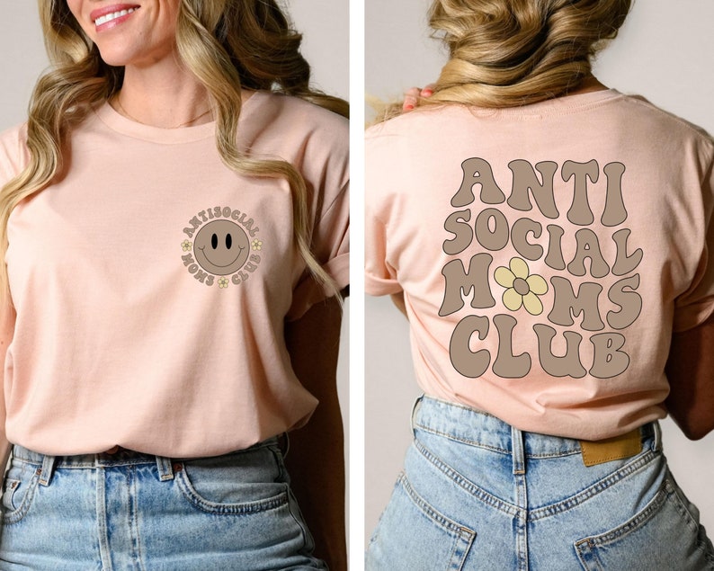 May include: A light pink t-shirt with a brown smiley face and the text "Antisocial Moms Club" on the front. The back of the shirt has the text "Antisocial Moms Club" in brown with a yellow flower.