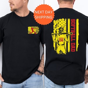 May include: Black t-shirt with a yellow and red softball player graphic on the front and a yellow and red American flag with the text "Softball Dad" on the back. The player is holding a bat and has the name "Emma" and number "06" on her jersey.