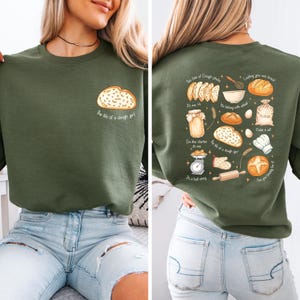 May include: An olive green sweatshirt with a bread-themed graphic on the front and back. The front features a bread loaf and the text "The life of a dough girl." The back has various baking-related illustrations and text.