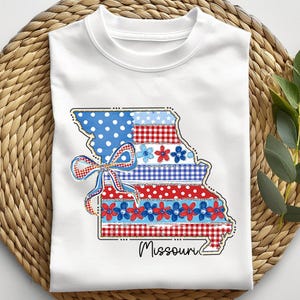 May include: White t-shirt featuring a colorful graphic of the Missouri state outline. The design incorporates red, white, and blue patterns, including polka dots, gingham, and floral elements, with a decorative bow. The word "Missouri" is written in script.