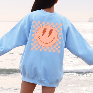May include: A light blue sweatshirt with a pink and orange chequered pattern and a smiley face with lightning bolts for eyes on the back.