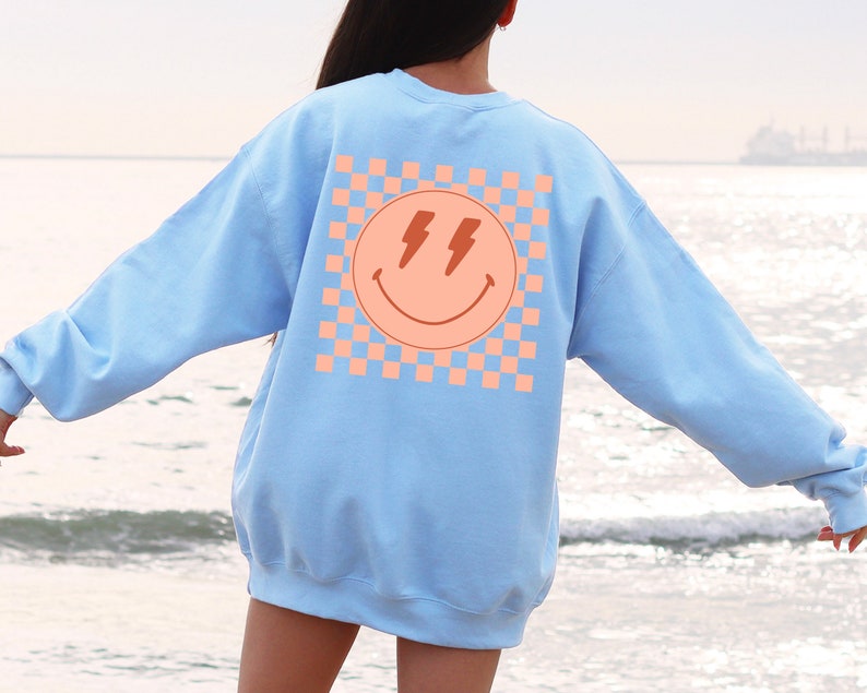 May include: A light blue sweatshirt with a pink and orange chequered pattern and a smiley face with lightning bolts for eyes on the back.