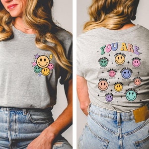 K&ouml;nnte beinhalten: Ein graues T-Shirt mit einem Motiv aus Smiley-Gesichtern und Sternen auf der Vorderseite und einem Motiv aus Smiley-Gesichtern mit inspirierenden Texten auf der R&uuml;ckseite. Der Text lautet "You Are" und enth&auml;lt S&auml;tze wie "Beautiful", "Victorious", "Strong", "Amazing", "Chosen", "Capable", "Always Loved" und "Never Alone".