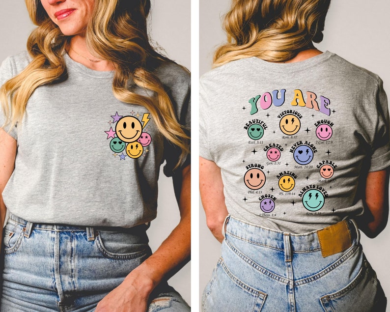 K&ouml;nnte beinhalten: Ein graues T-Shirt mit einem Motiv aus Smiley-Gesichtern und Sternen auf der Vorderseite und einem Motiv aus Smiley-Gesichtern mit inspirierenden Texten auf der R&uuml;ckseite. Der Text lautet "You Are" und enth&auml;lt S&auml;tze wie "Beautiful", "Victorious", "Strong", "Amazing", "Chosen", "Capable", "Always Loved" und "Never Alone".