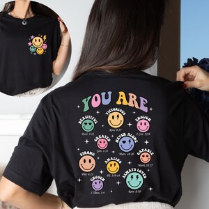 K&ouml;nnte beinhalten: Schwarzes T-Shirt mit einer bunten Grafik auf der R&uuml;ckseite, die "You Are" mit einem Smiley f&uuml;r jede positive Affirmation sagt. Die Affirmationen beinhalten: Beautiful, Victorious, Created, Strong, Chosen, Amazing, Never Alone, Enough, Capable, Always Loved. Jede Affirmation enth&auml;lt einen Bibelvers.