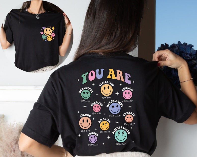 K&ouml;nnte beinhalten: Schwarzes T-Shirt mit einer bunten Grafik auf der R&uuml;ckseite, die "You Are" mit einem Smiley f&uuml;r jede positive Affirmation sagt. Die Affirmationen beinhalten: Beautiful, Victorious, Created, Strong, Chosen, Amazing, Never Alone, Enough, Capable, Always Loved. Jede Affirmation enth&auml;lt einen Bibelvers.