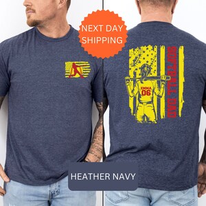May include: A heather navy blue t-shirt with a softball player graphic on the back. The graphic features a woman holding a bat in front of an American flag with the text "Softball Dad". The front of the shirt has a smaller graphic of a softball player.