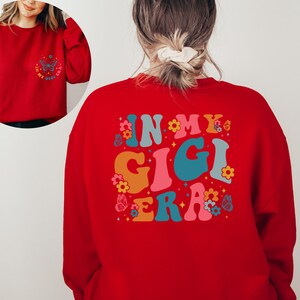 May include: Red sweatshirt with the text "In My Gigi Era" in colorful retro font with butterflies and flowers.