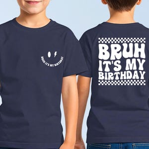 May include: A navy blue t-shirt with a white smiley face and the text "Bruh it's my birthday" on the front. The back of the shirt has the text "Bruh it's my birthday" in a white checkered box.